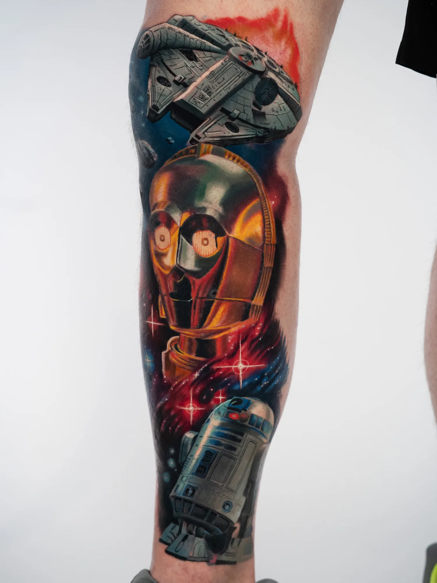 Full color Star Wars tattoo on a lower leg showing the Millennium Falcon, a C-3PO portrait and R2-D2 against a red and blue nebula.