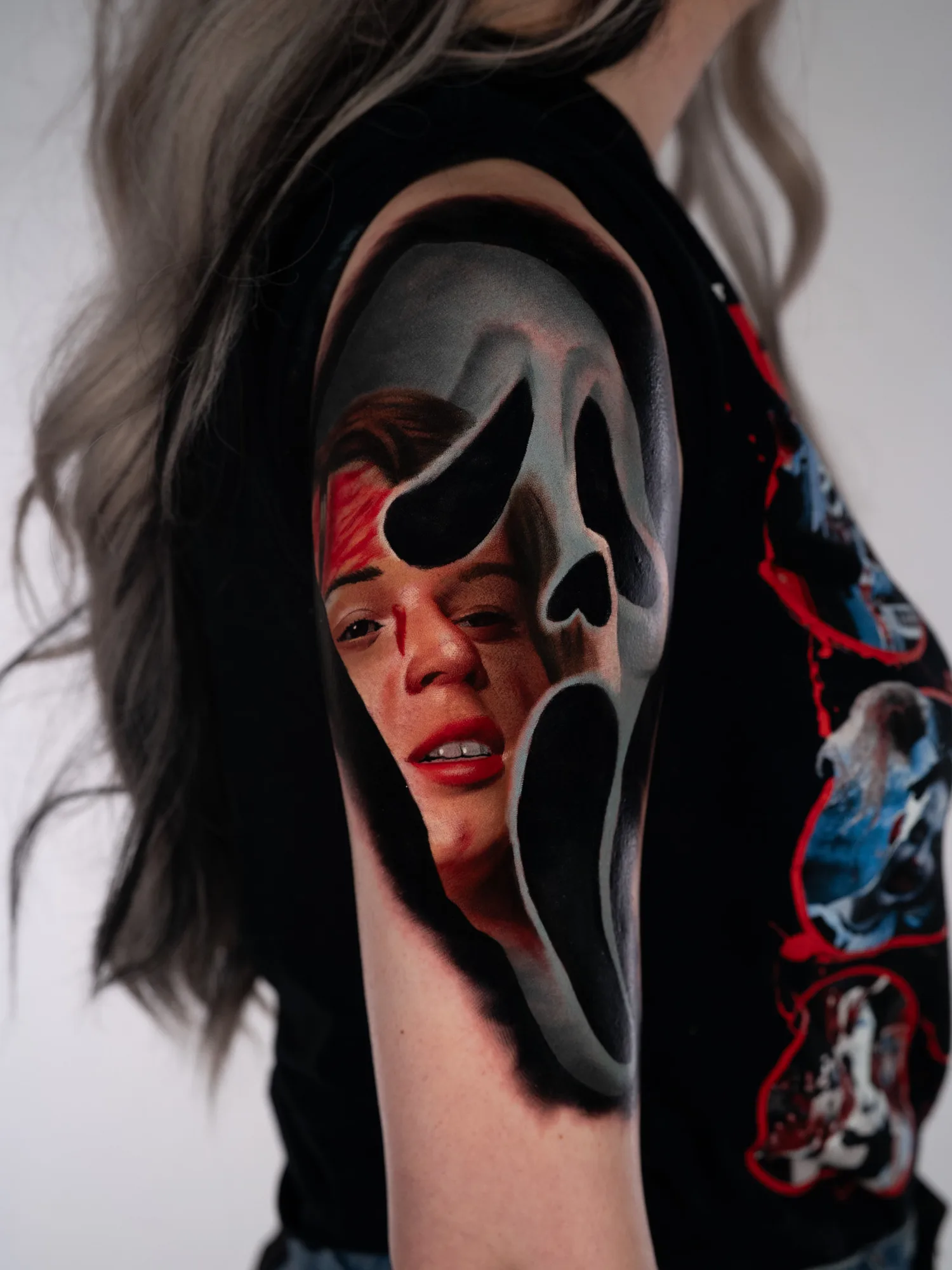 Color realism tattoo of a Ghostface mask on an upper arm, with a screaming woman's face rendered inside the mask's opening.