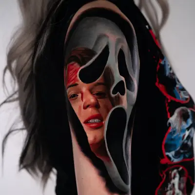 Color realism tattoo of a Ghostface mask on an upper arm, with a screaming woman's face rendered inside the mask's opening.