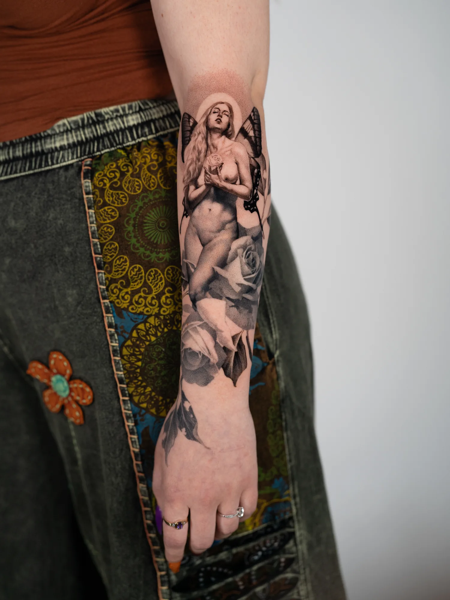 Black and grey realism tattoo of a nude winged fairy holding a rose, above two large roses, running down an inner forearm to the wrist.