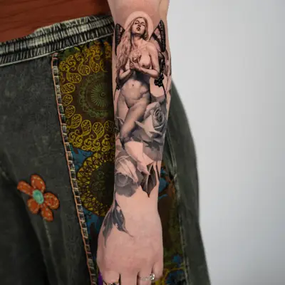 Black and grey realism tattoo of a nude winged fairy holding a rose, above two large roses, running down an inner forearm to the wrist.