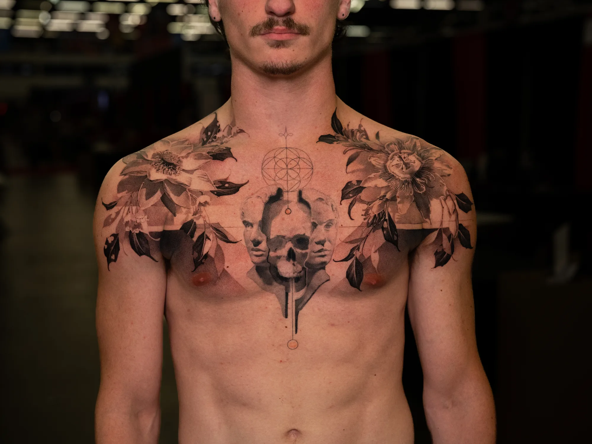 Black and grey chest tattoo with a passionflower across each shoulder, a skull flanked by two classical faces at the center, and fine line geometric work above it.
