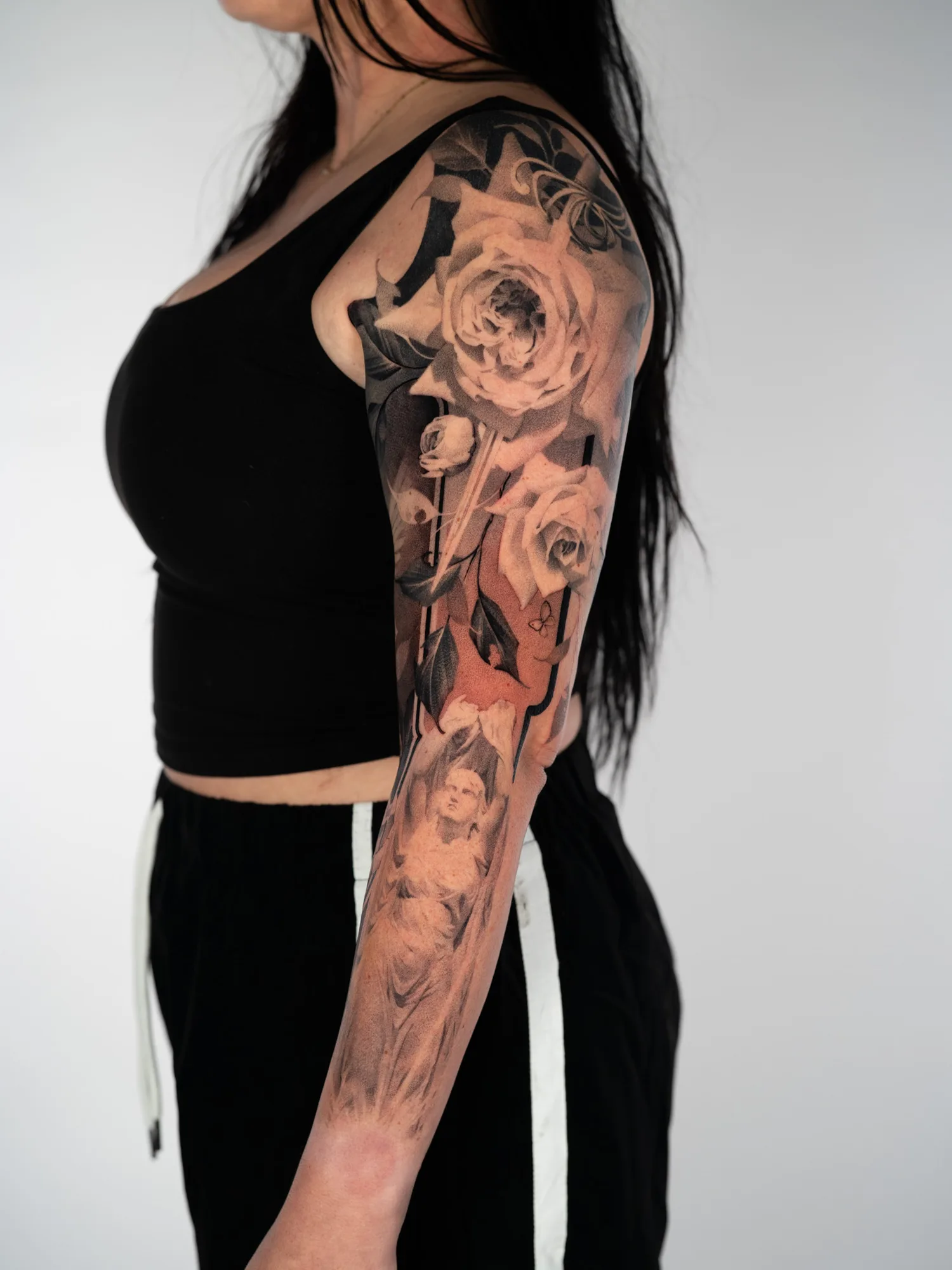 Black and grey realism full sleeve with large roses on the upper arm, a sword blade running down through them, and a draped classical statue on the forearm.