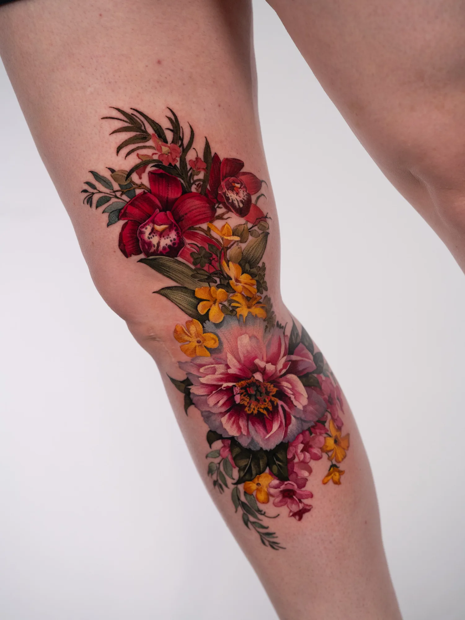 Full color realism tattoo of deep red orchids, yellow and orange flowers and a large pink peony running down the inside of a leg across the knee.