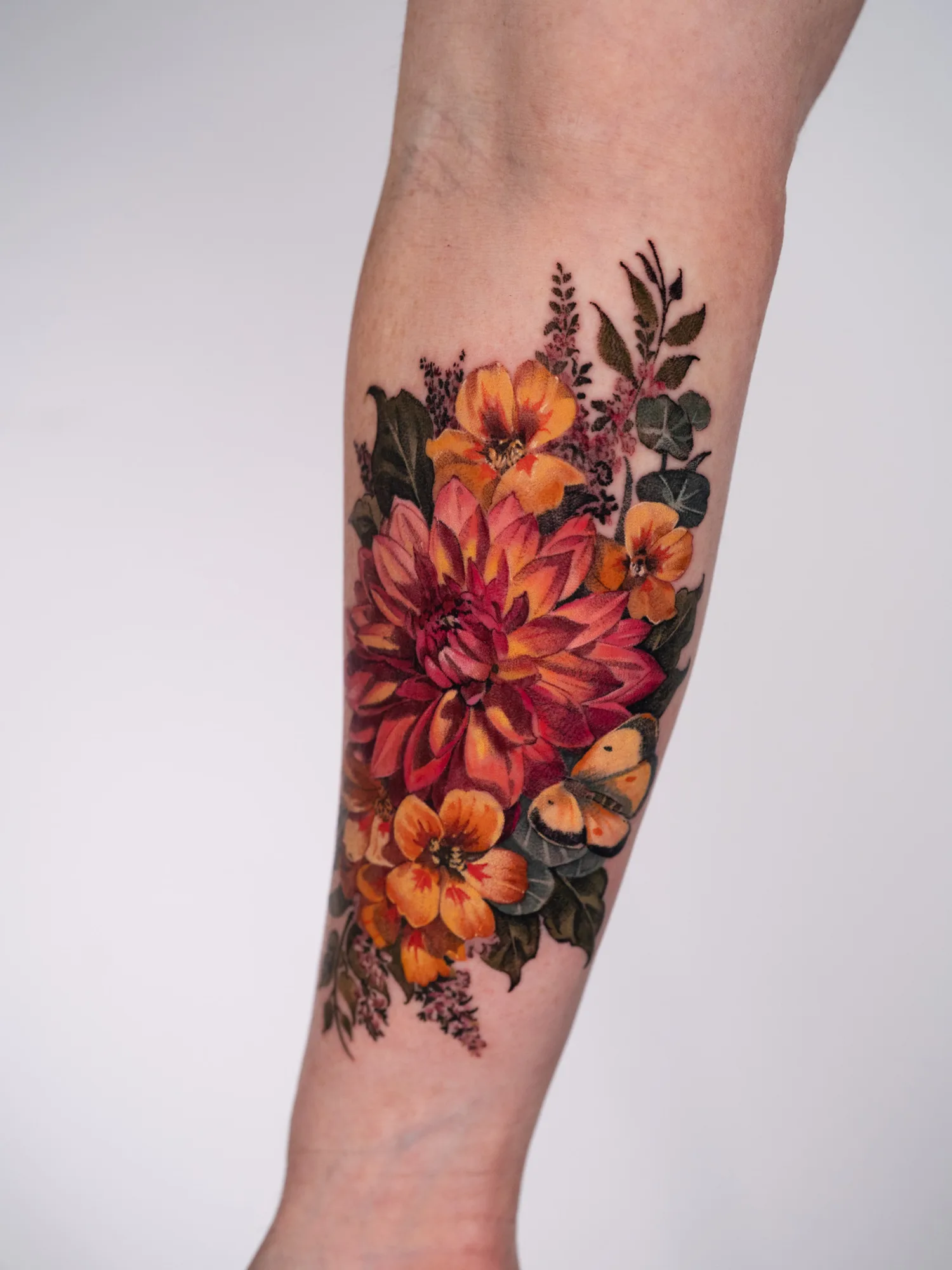 Full color realism tattoo of a red and orange dahlia with orange blooms, heather sprigs, eucalyptus and a yellow butterfly, on an inner forearm.
