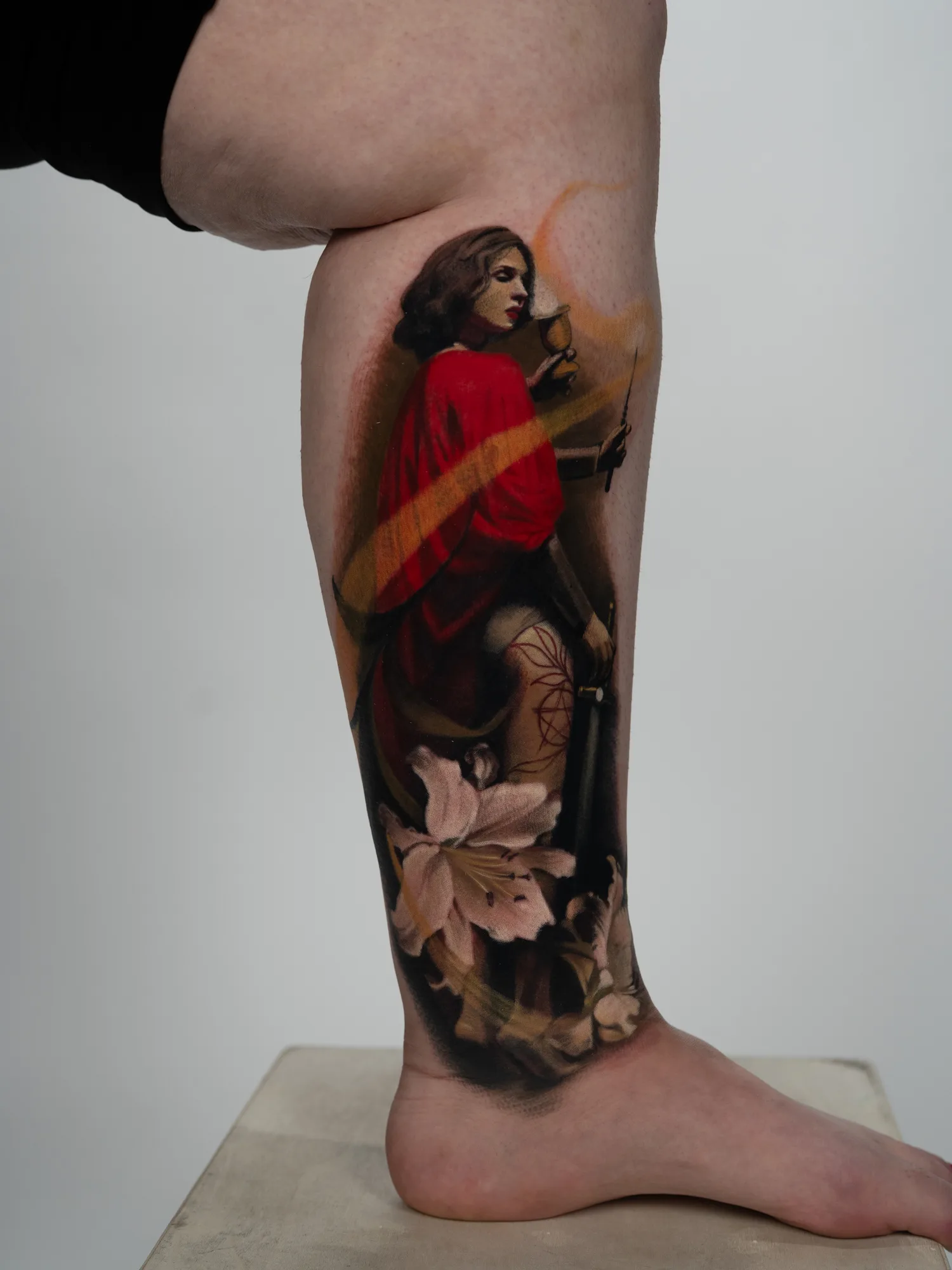 Tattoo of a woman in a red robe, with a white lily on a woman's calf.