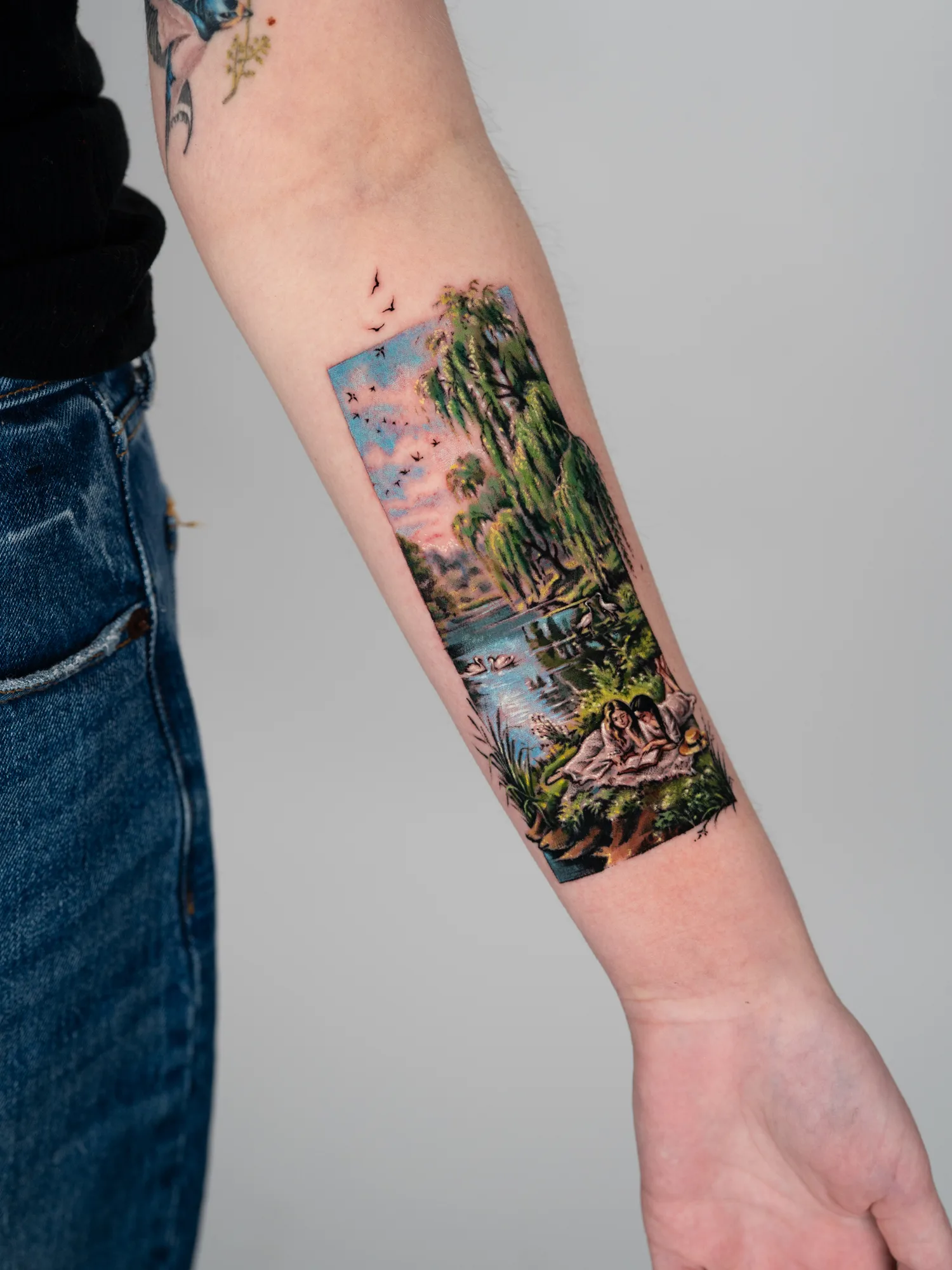 Landscape tattoo on the forearm in color of two women lying in the grass.