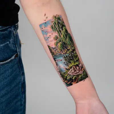 Landscape tattoo on the forearm in color of two women lying in the grass.