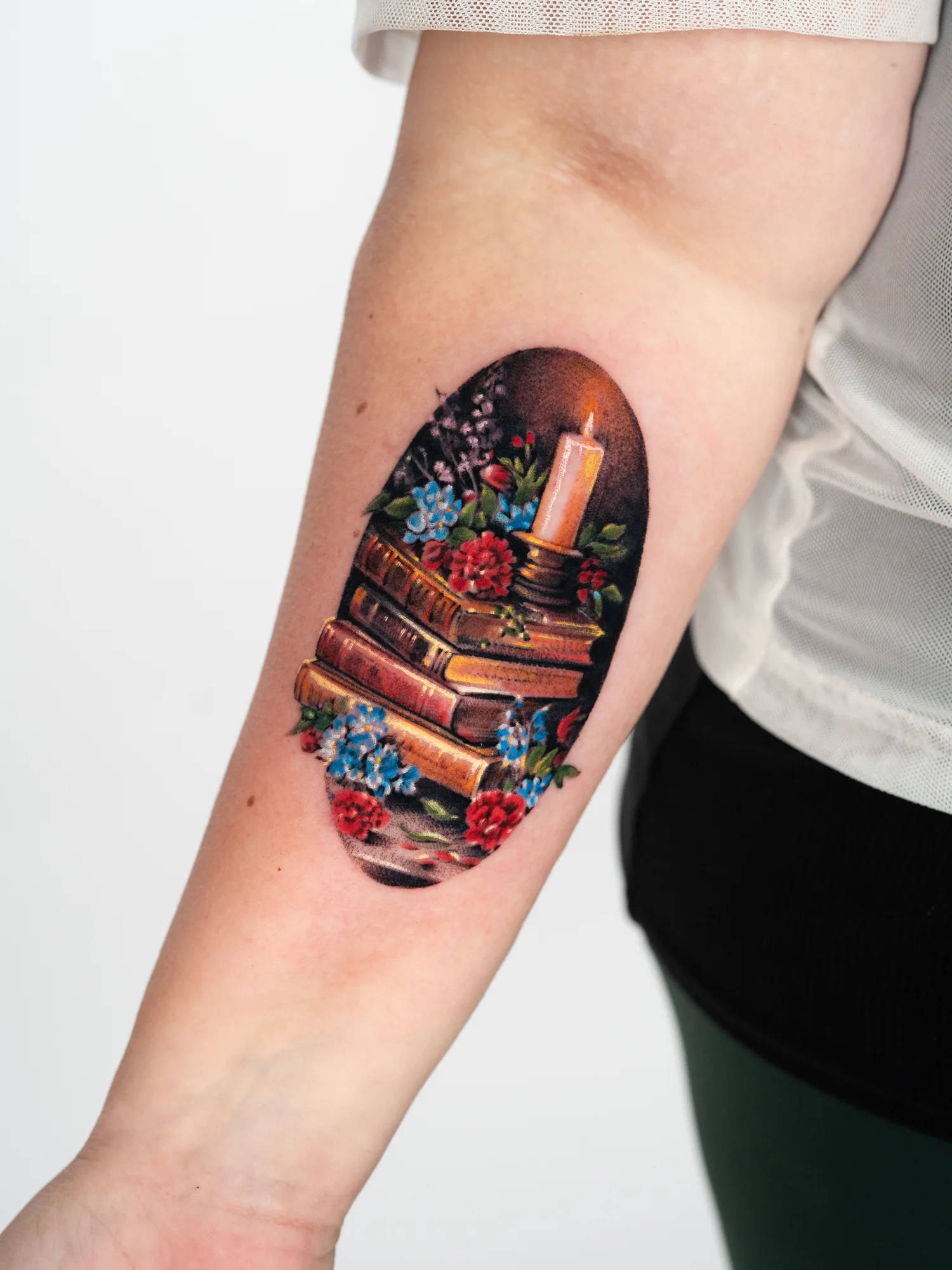 Tattoo of a still life with books, a candle, and flowers on an inner forearm