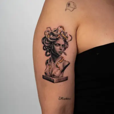Tattoo of a bust of Medusa on an upper forearm in black and grey with gold snakes.