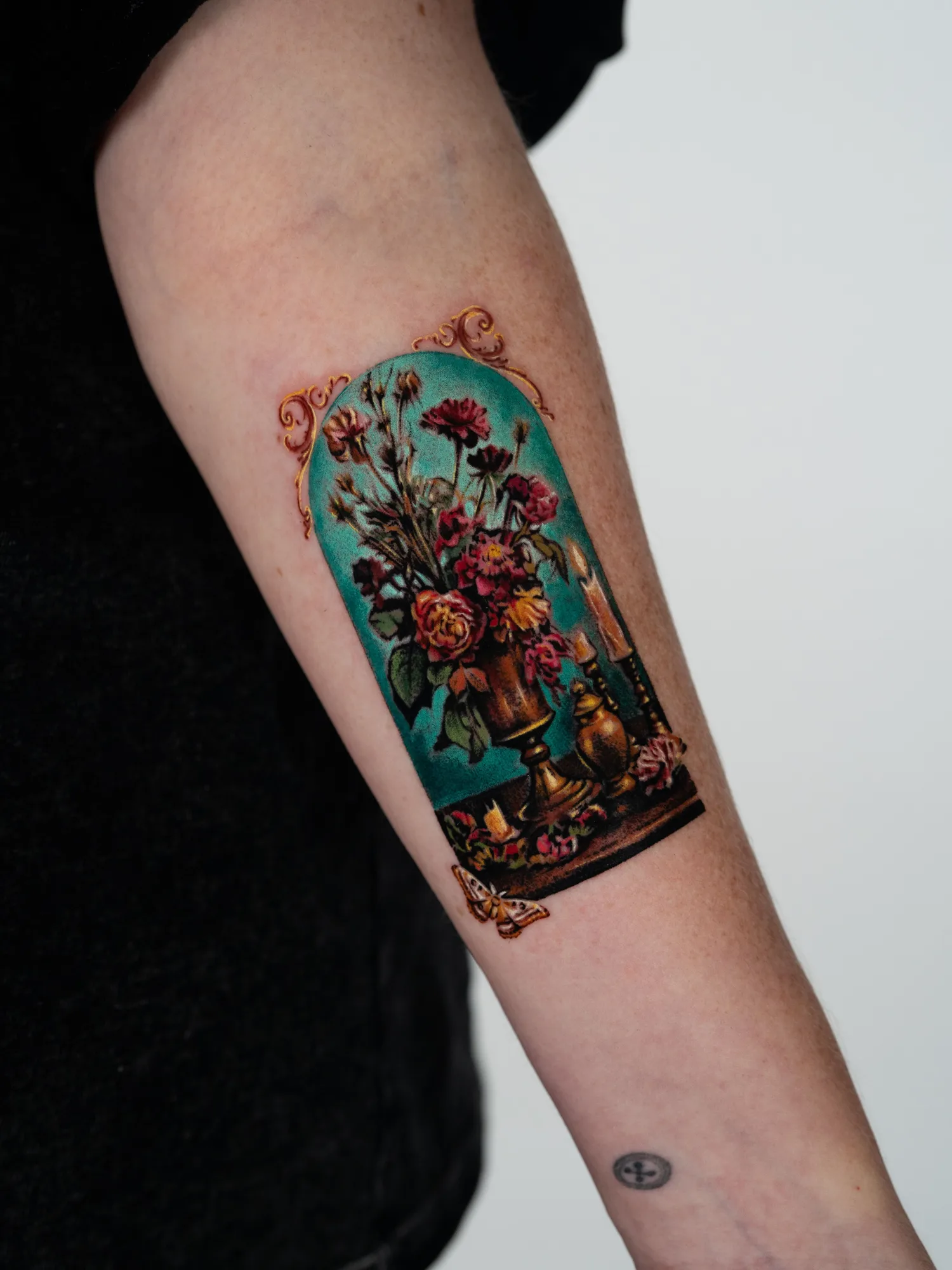 Color microrealism tattoo of a decaying floral still life in an arched frame, with a brass urn, a burning candle and a moth, on a forearm.