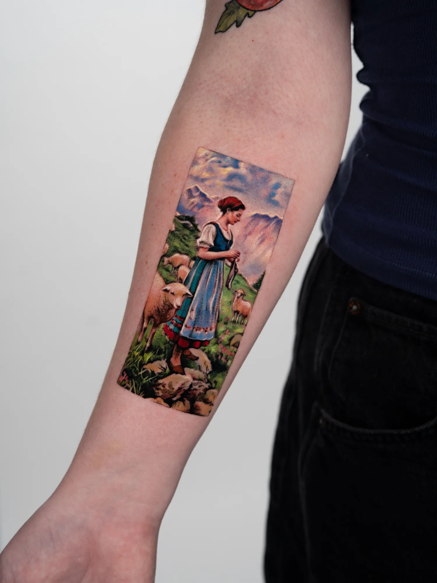 Color painterly tattoo of a shepherdess in a dirndl standing among sheep in an alpine meadow with snow-capped mountains behind, on a forearm.