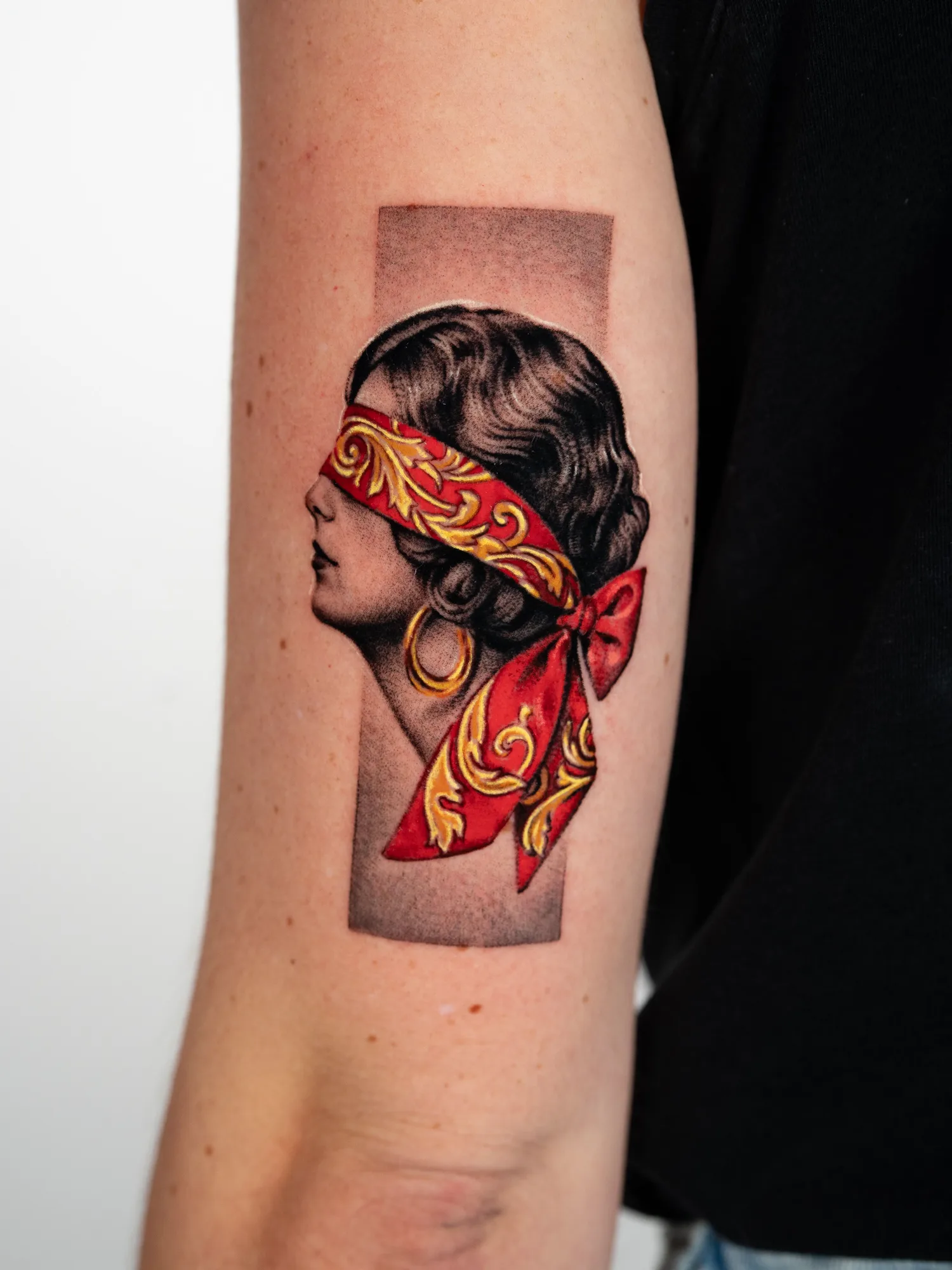 Black and grey tattoo of a woman's profile blindfolded with a red silk band painted in gold filigree and tied in a bow, on the back of an arm.