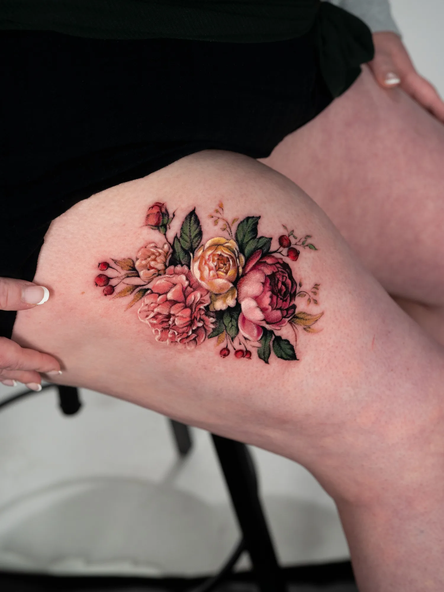 Color realism tattoo of a loose bundle of roses, peonies and red berries with no frame or background, on a thigh.