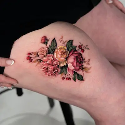 Color realism tattoo of a loose bundle of roses, peonies and red berries with no frame or background, on a thigh.