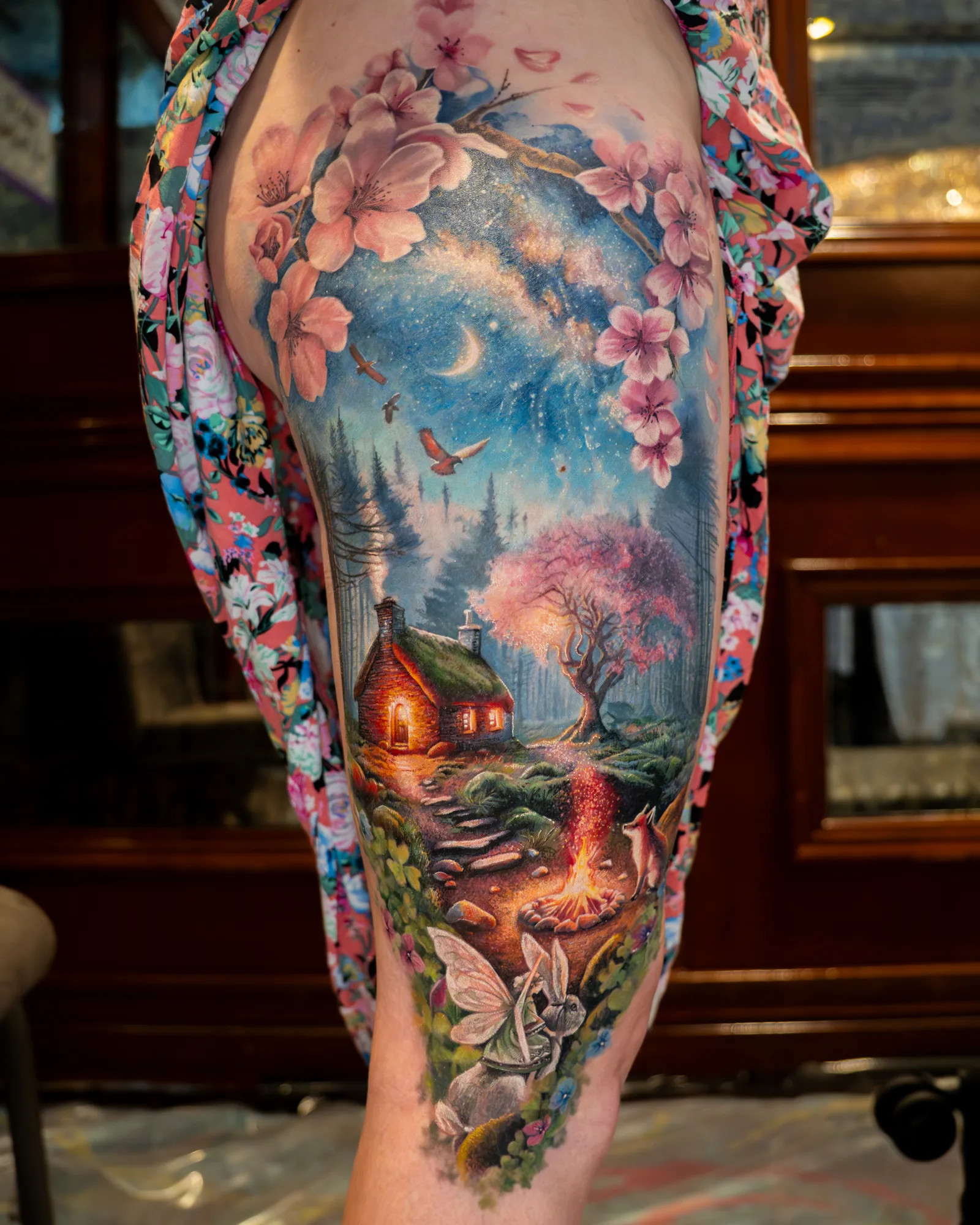 Colorful tattoo depicting a whimsical landscape with a cabin, cherry blossoms, and animals.