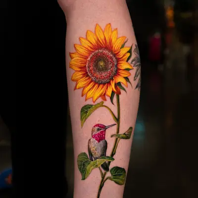 Tattoo of a sunflower and hummingbird on a person's leg.