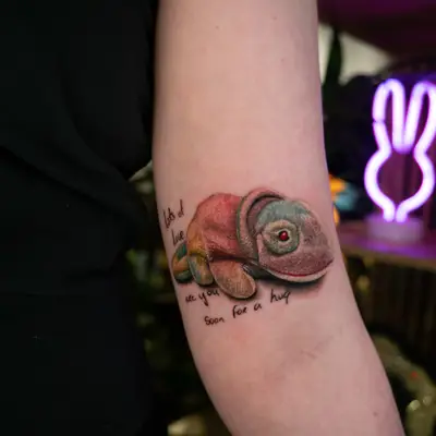 A realistic, colorful chameleon tattoo with text on an arm.