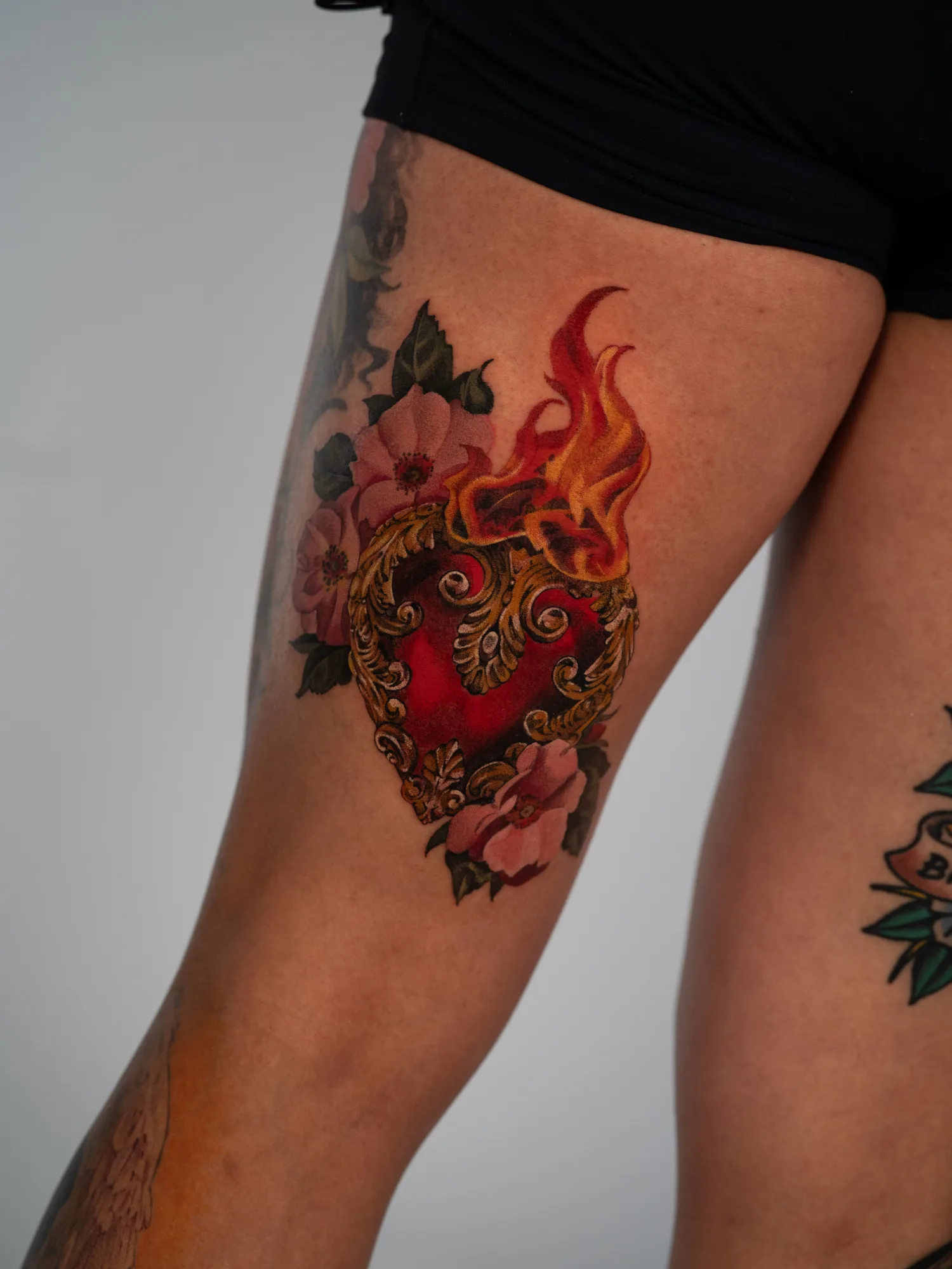 Tattoo of a sacred heart on a thigh surrounded by flowers and flames.