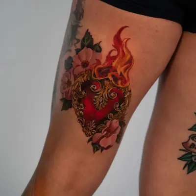 Tattoo of a sacred heart on a thigh surrounded by flowers and flames.