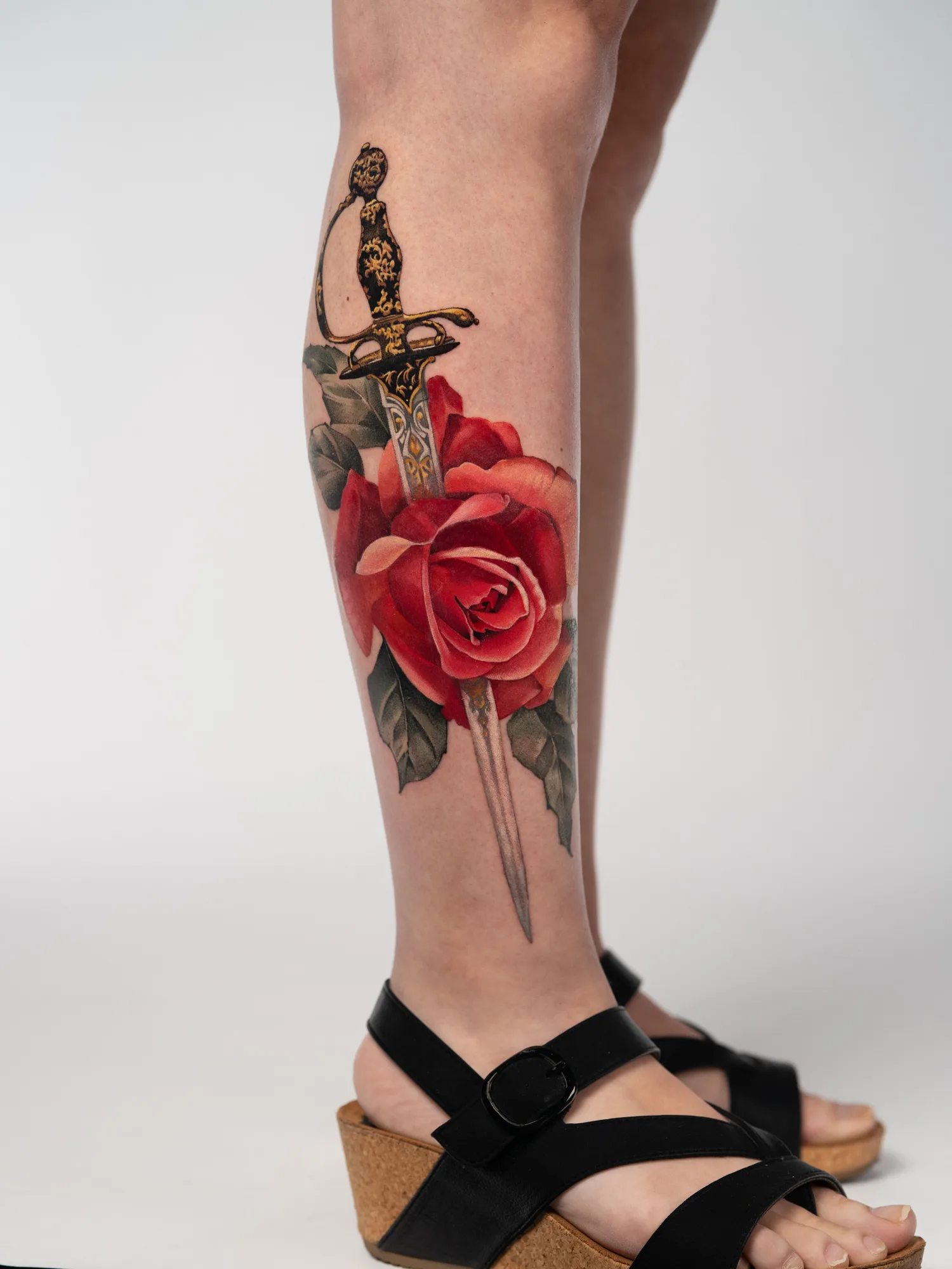 Color realism tattoo of a red rose with an ornate dagger piercing through it on the calf.