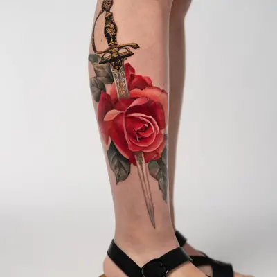 Color realism tattoo of a red rose with an ornate dagger piercing through it on the calf.