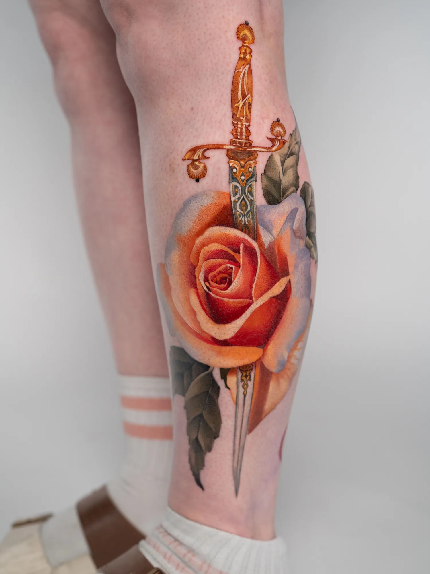 Color realism tattoo of a peachy-white rose with an ornate dagger piercing through it on the calf.