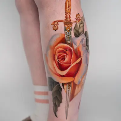 Color realism tattoo of a peachy-white rose with an ornate dagger piercing through it on the calf.