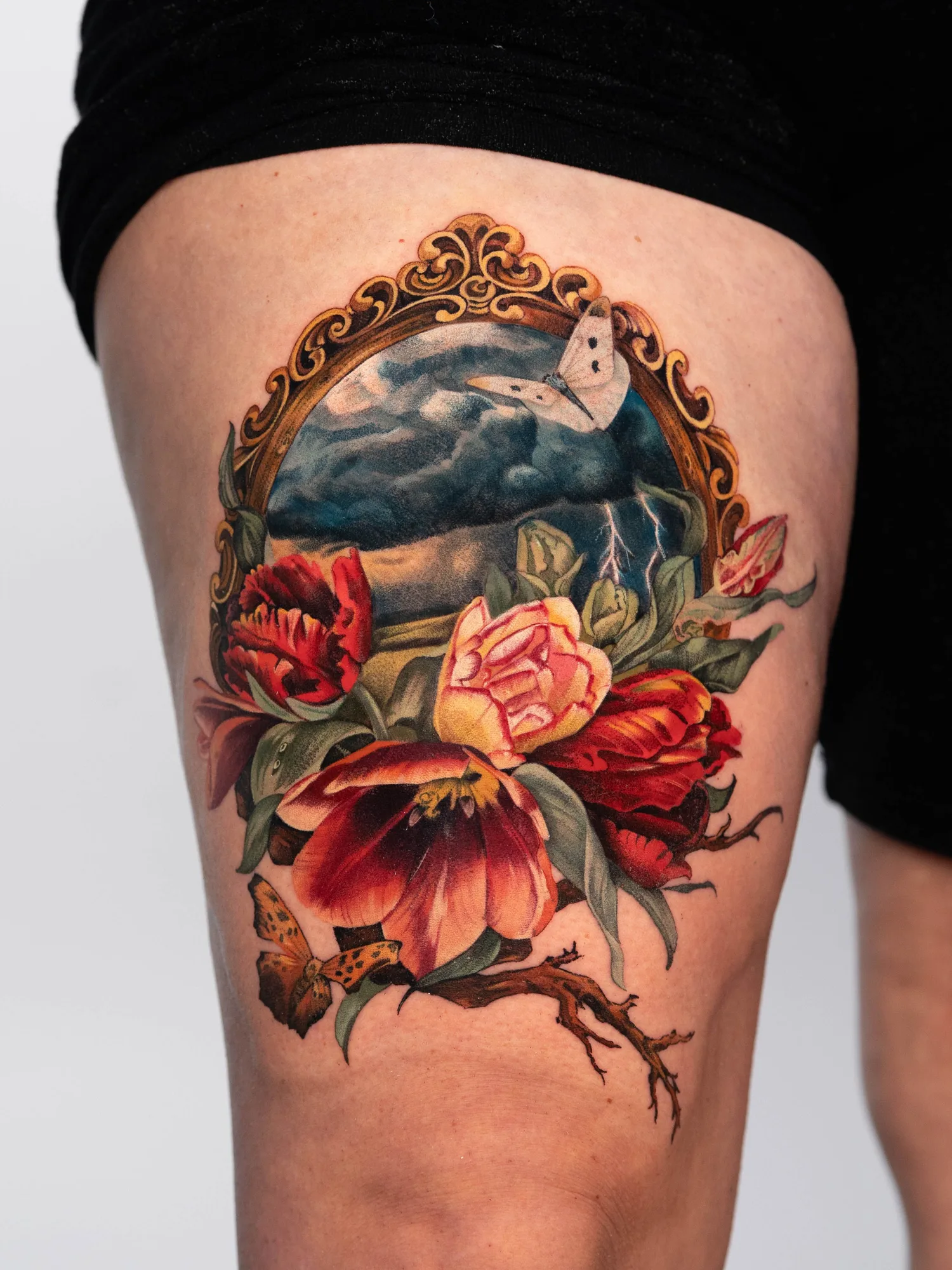 Vibrant floral tattoo with a butterfly and landscape within a gold decorative frame.