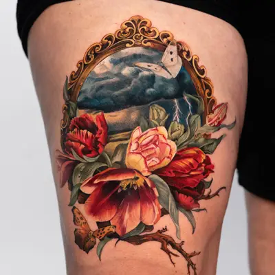 Vibrant floral tattoo with a butterfly and landscape within a gold decorative frame.