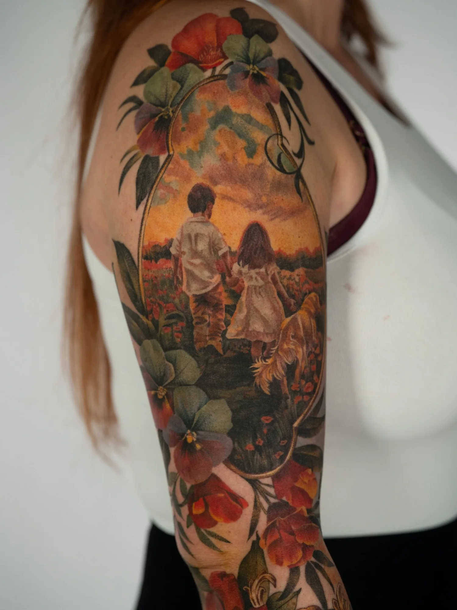 Landscape tattoo of a boy and a girl with a dog in a field during a sunset in a gold frame with flowers