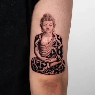 Black and grey tattoo of a seated Buddha with patterned robe on the upper arm.