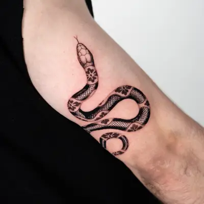 Tattoo of a patterned snake on an inner bicep in black and grey.