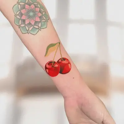 Tattoo of two red cherries with a green leaf on an inner forearm.
