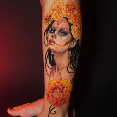 A colorful portrait tattoo featuring a woman with marigolds and Dia de los Muertos makeup on a leg.