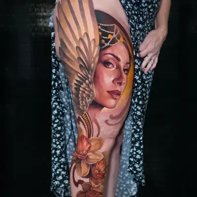 Color tattoo running from the hip to below the knee on the outside of a leg: a realistic portrait of a woman in a beaded headdress with a large golden wing behind her, and orange daffodils with Art Nouveau swirls below.