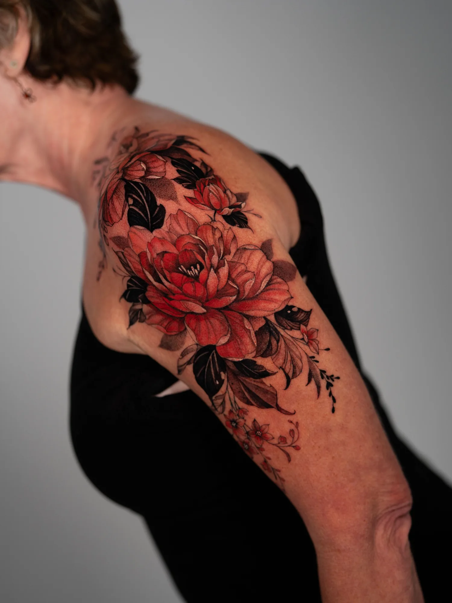 Colorful floral tattoo on woman's shoulder and arm featuring red flowers and black leaves in a fineline style.