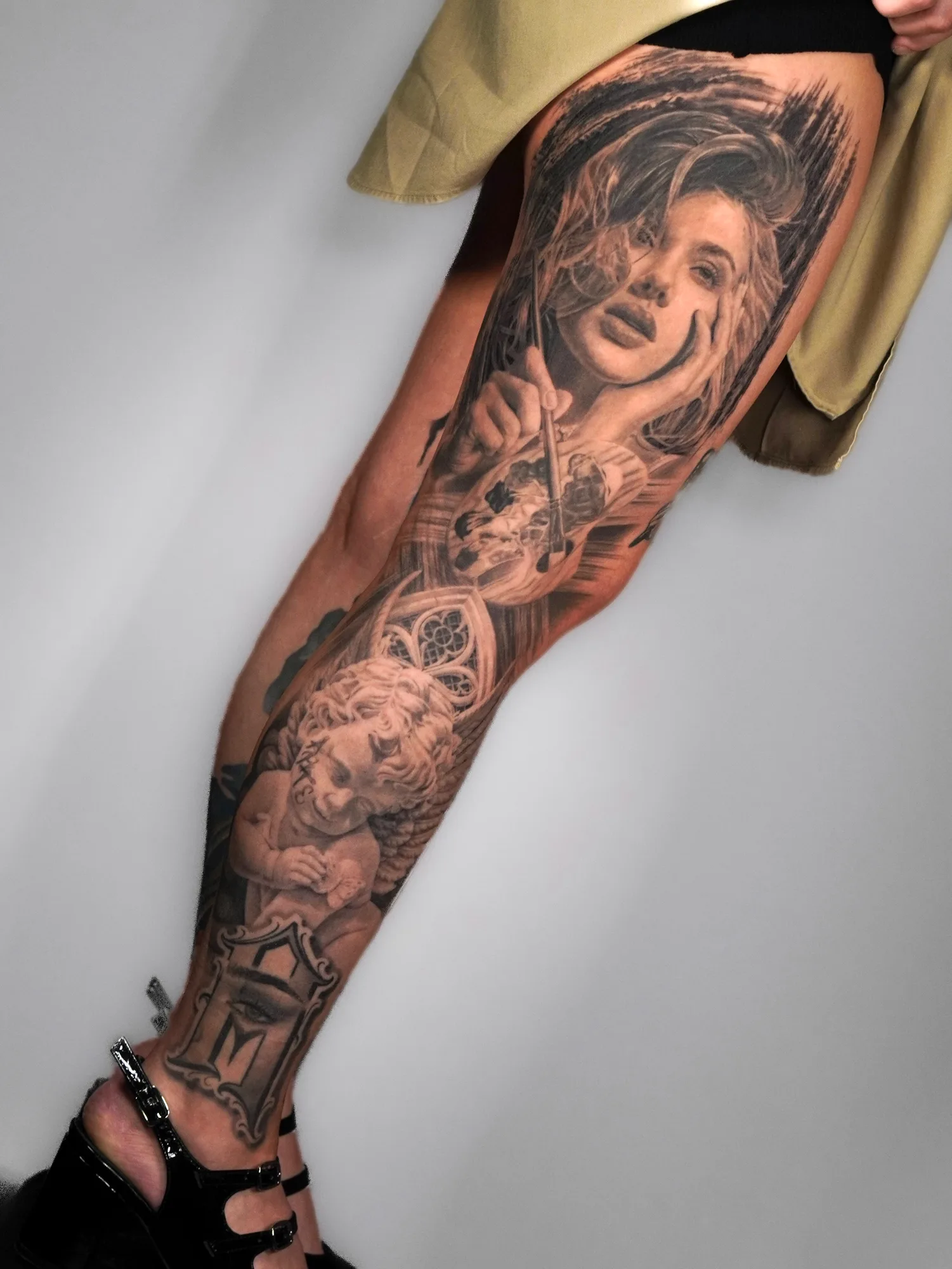 Leg tattoo depicting a woman's face and cherub in black and grey realism style.