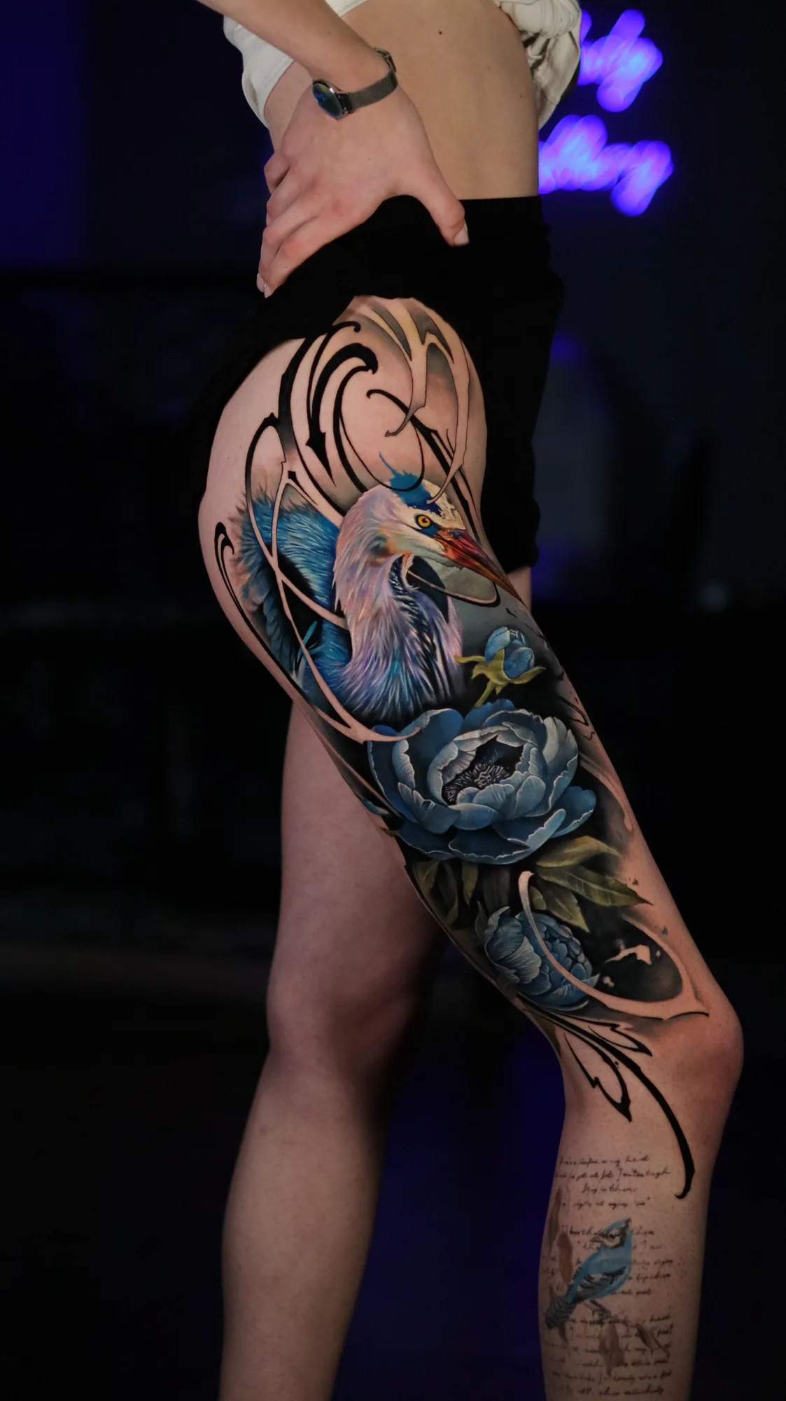 Colorful blue heron and floral tattoo on the thigh with calligraphy elements..
