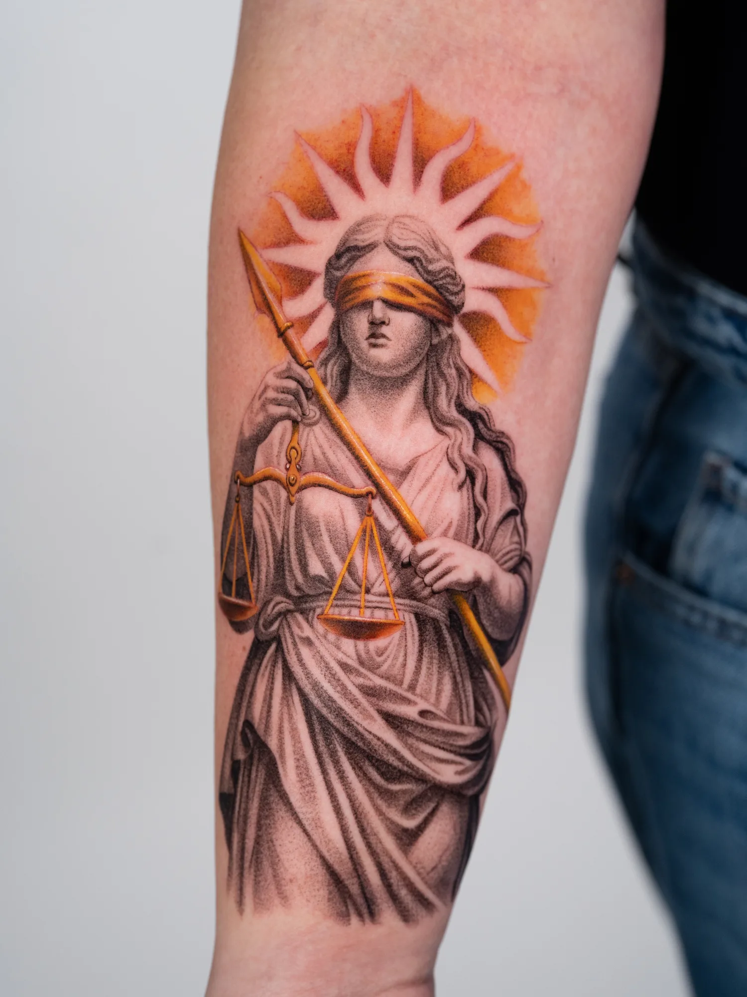 Blindfolded Lady Justice tattoo with scales and sunburst on arm.