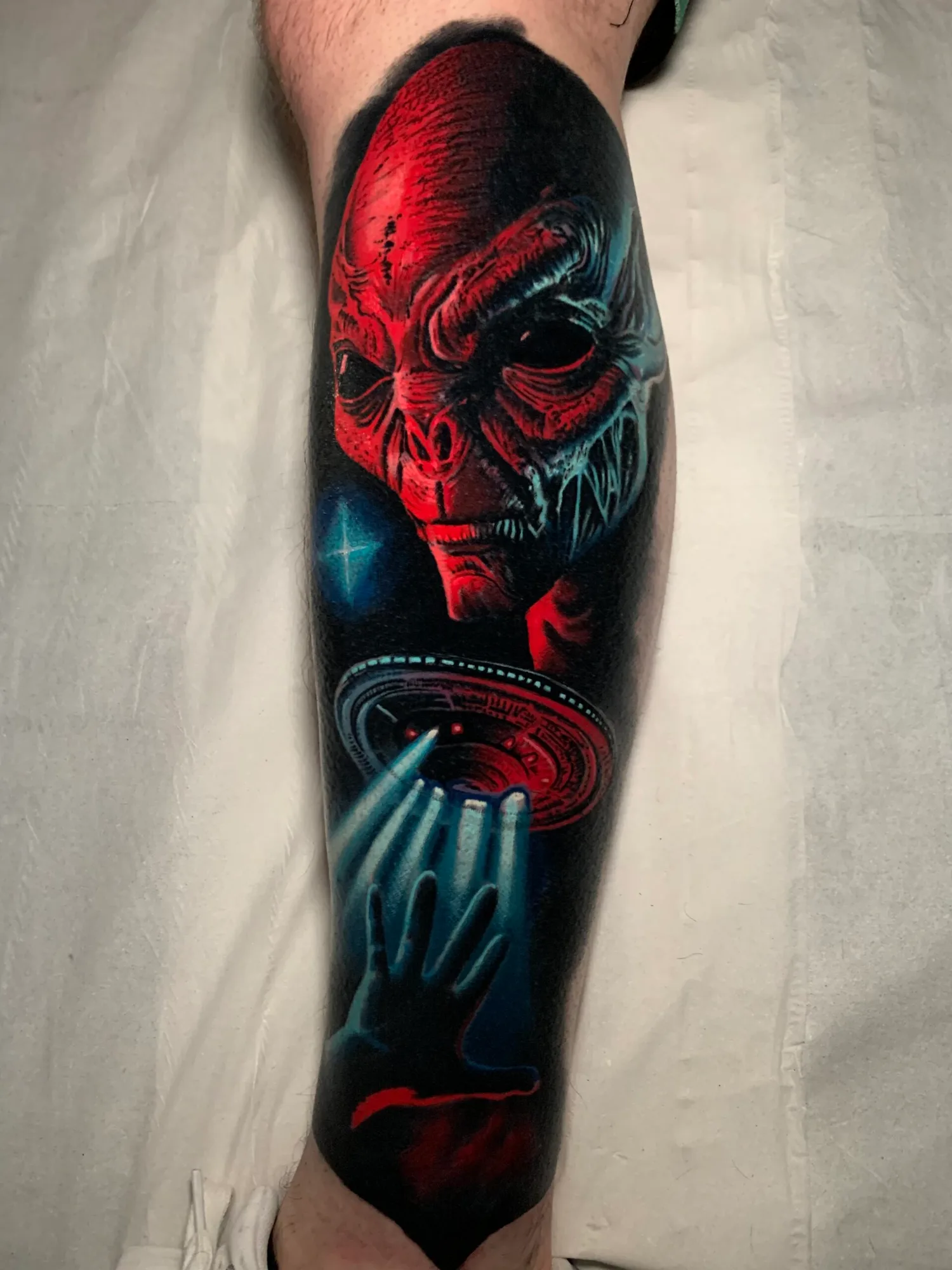 Tattoo depicting an alien face with a UFO and hand reaching towards light.