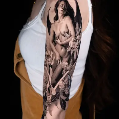 Black and grey tattoo of nude figure with wings and flowers on arm