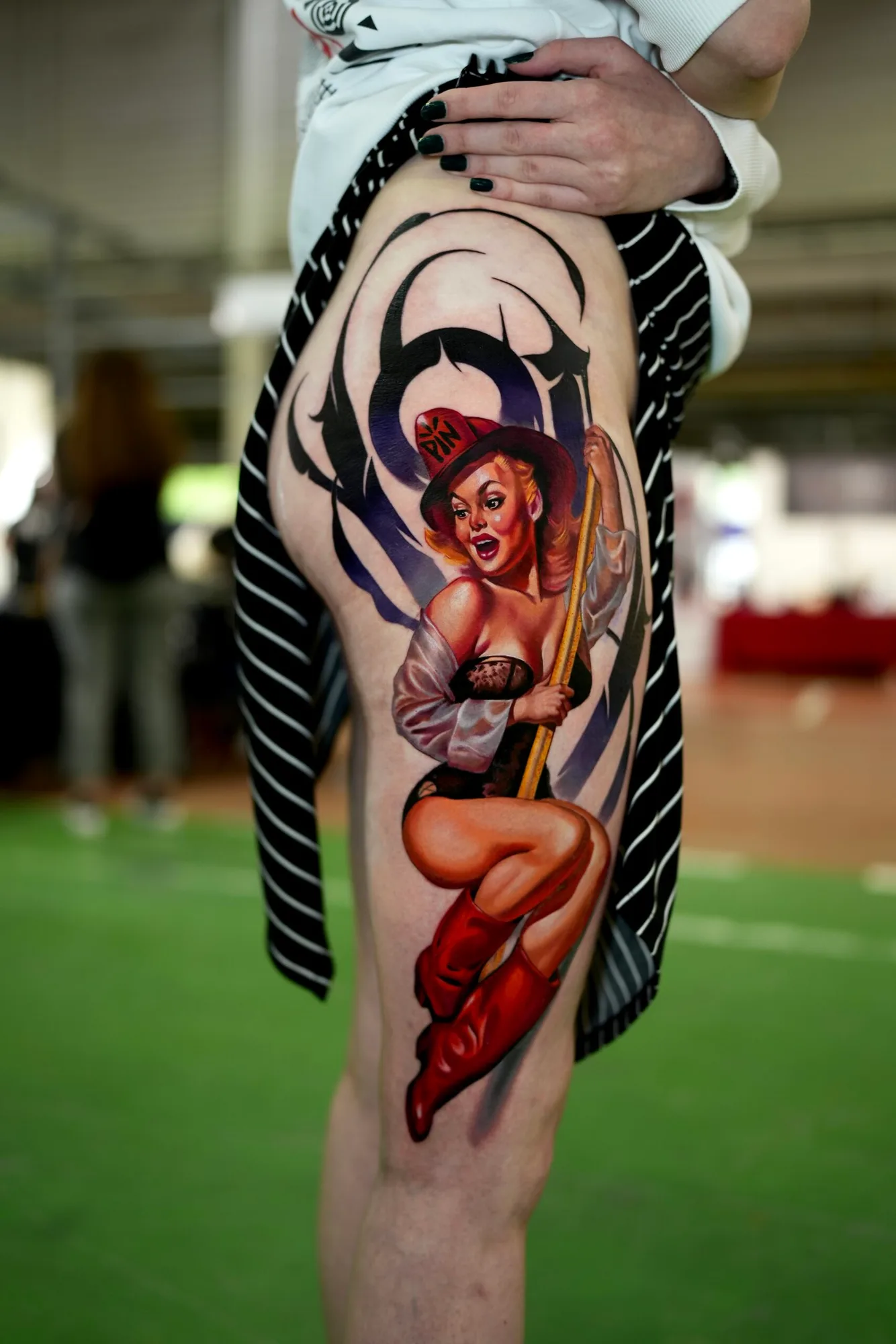 Pin-up style tattoo of a woman on a pole, wearing red boots and hat, on thigh.