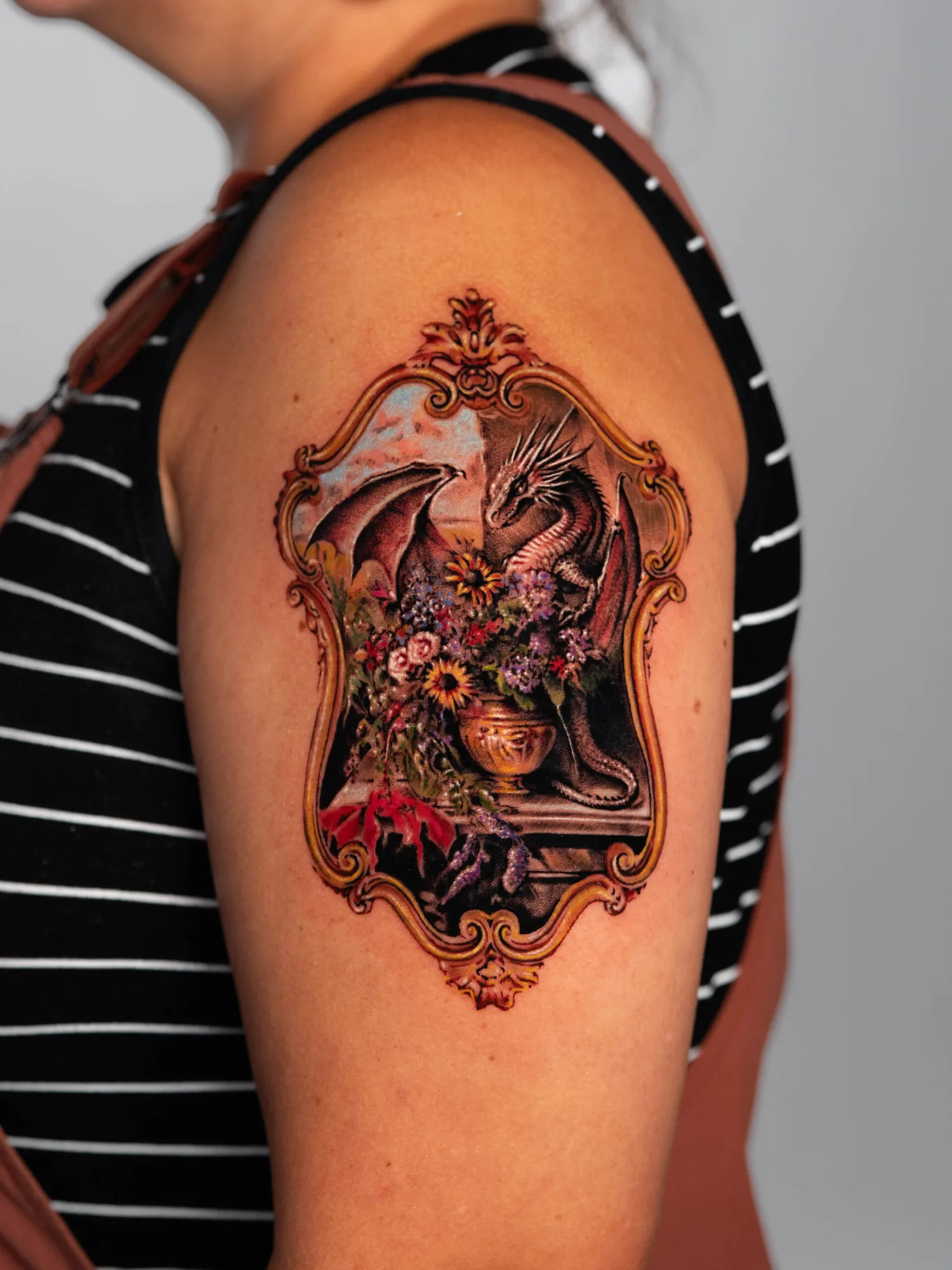 Tattoo of a dragon with flowers in a decorative frame on upper arm.