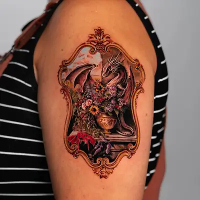 Tattoo of a dragon with flowers in a decorative frame on upper arm.