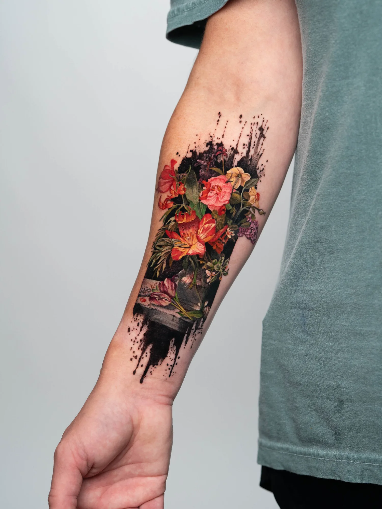Forearm tattoo of colorful flowers with black ink splashes.