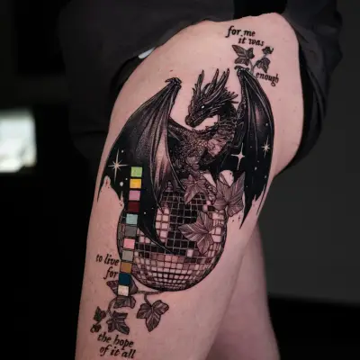 Tattoo of dragon on disco ball with text and leaves on thigh.