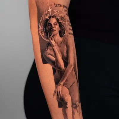 Intricate black and grey tattoo of a woman surrounded by text and floral design on the forearm.