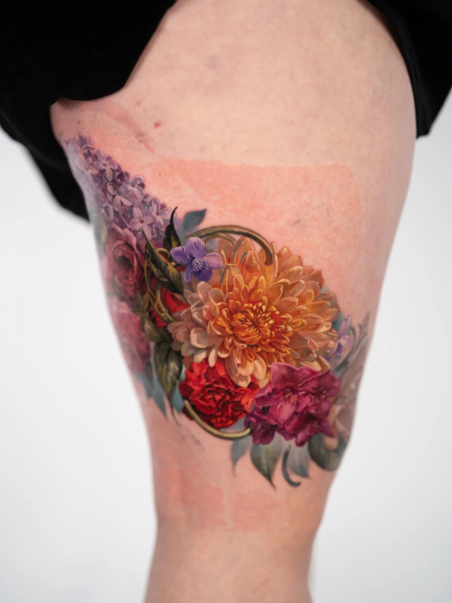 Color realism tattoo of lilacs, a wood violet, an orange chrysanthemum, a red carnation and a magenta rhododendron with gold scrollwork, on an outer right thigh.