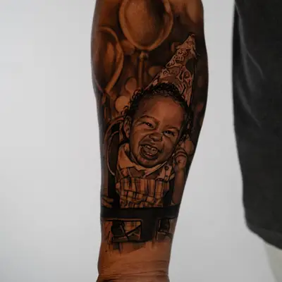 Black and grey realism tattoo of a laughing toddler in a paper party hat sitting in a high chair, with balloons behind, running down a forearm.